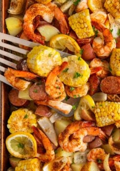 A sheet pan shrimp boil filled with seasoned baked shrimp, corn on the cob slices, red potatoes, sausage, lemon wedges, and chopped herbs, with a spatula lifting a serving.