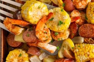 A sheet pan shrimp boil filled with seasoned baked shrimp, corn on the cob slices, red potatoes, sausage, lemon wedges, and chopped herbs, with a spatula lifting a serving.