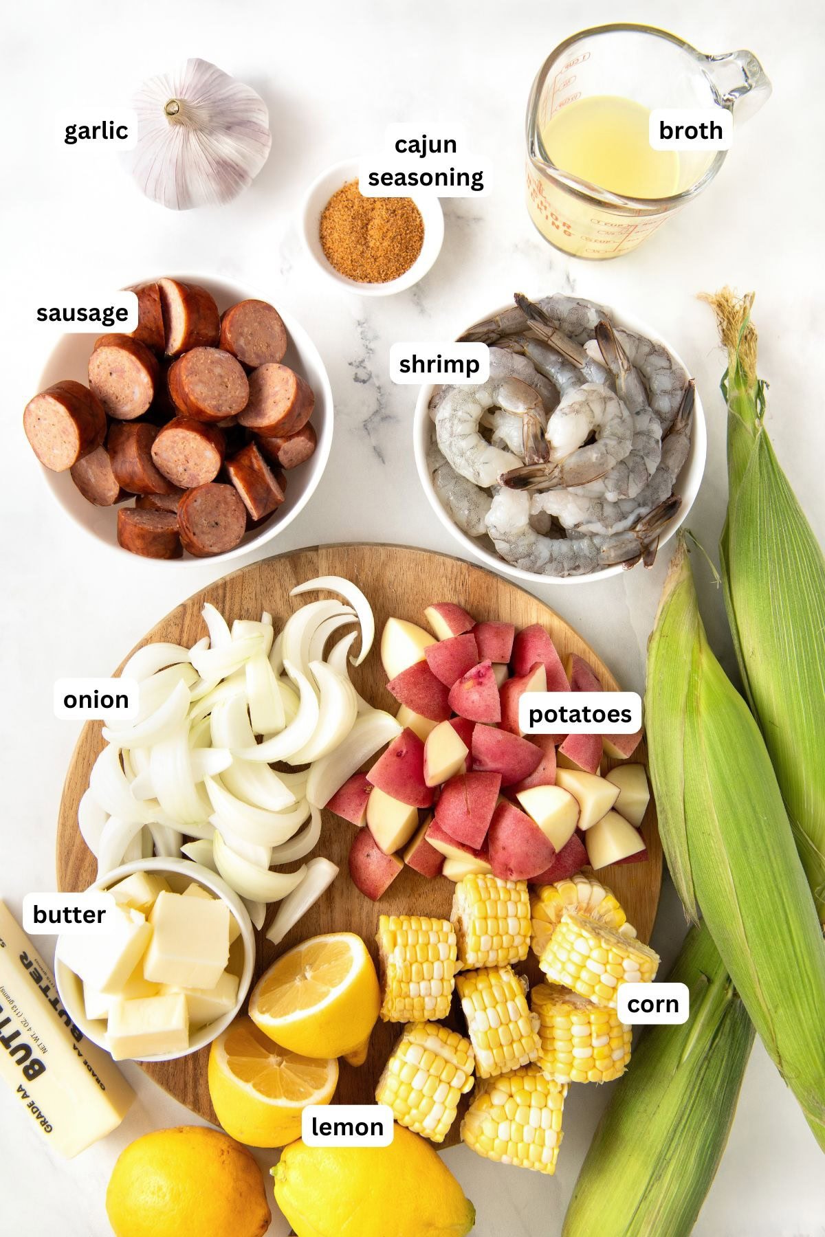 Ingredients for a Cajun sheet pan shrimp boil recipe are arranged on a white surface, including labeled bowls of sausage, shrimp, broth, cajun seasoning, onion, potatoes, corn, garlic, butter, and lemon, plus two ears of corn and whole lemons.