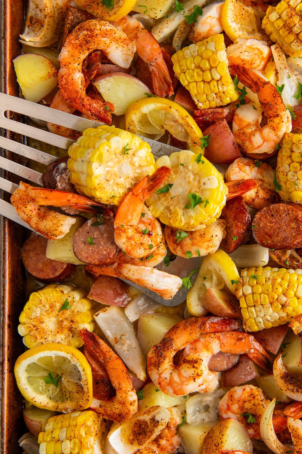 Sheet Pan Shrimp Boil features juicy shrimp, smoky sausage, tender potatoes and sweet corn all roasted in a cajun seasoned garlic butter. A sheet pan shrimp boil filled with seasoned baked shrimp, corn on the cob slices, red potatoes, sausage, lemon wedges, and chopped herbs, with a spatula lifting a serving.