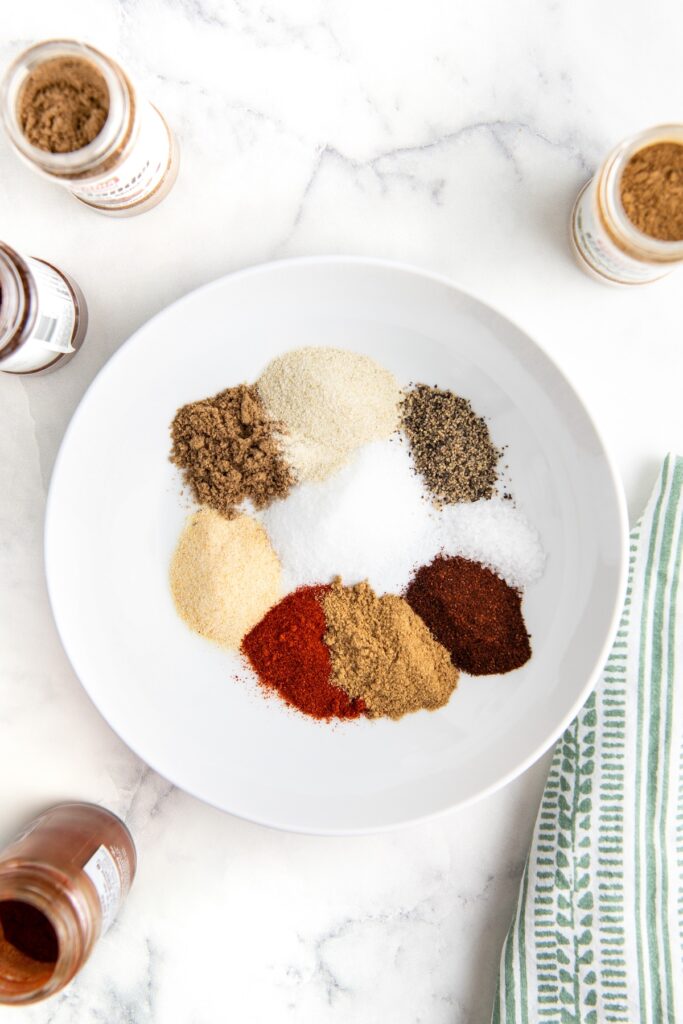 A white plate holds assorted ground spices arranged in small piles, including garlic, onion powder, sea salt, paprika, coriander, cumin, chili powder, black pepper, and Accent seasoning. Spice jars surround the plate, and a green striped cloth is partially visible on the right.