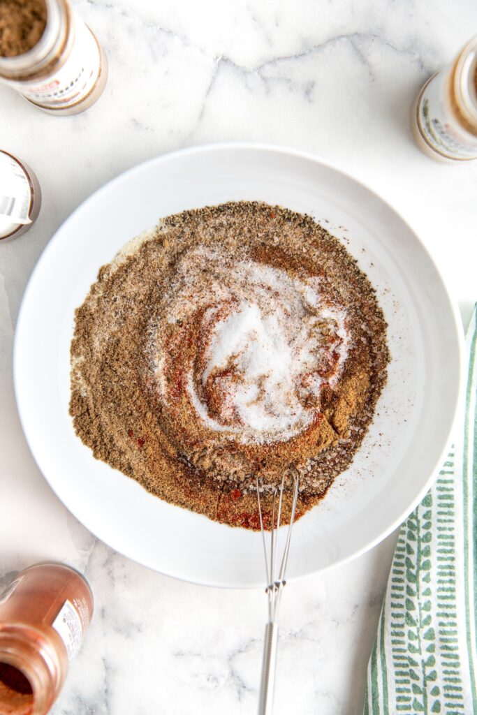 A white plate on a marble surface holds a mix of ground spices and salt swirling together, with a small whisk resting inside. Spice jars and a green-striped cloth are nearby.