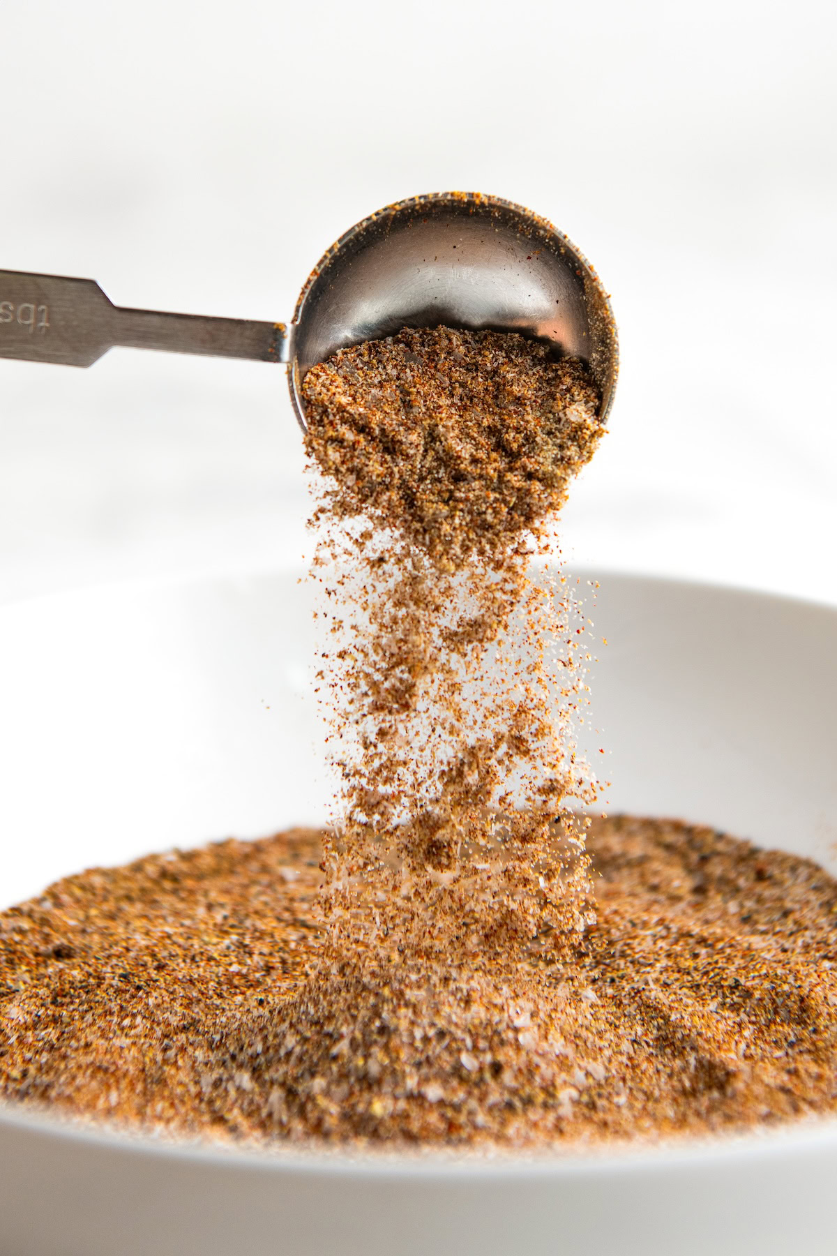 A metal measuring spoon pours a blend of authentic fajita seasoning mix into a white bowl filled with the same spice mixture.