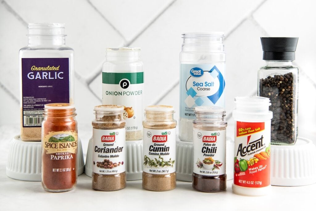 A selection of spice containers for making authentic fajita seasoning recipe, including granulated garlic, onion powder, sea salt, black peppercorns, paprika, coriander, cumin, chili powder, and Accent seasoning.