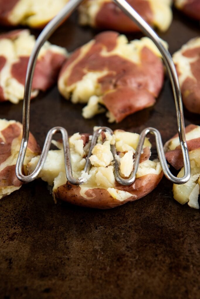 A metal potato masher presses down on a small red potato, partially smashing it on a dark surface with other partially smashed red potatoes in the background.
