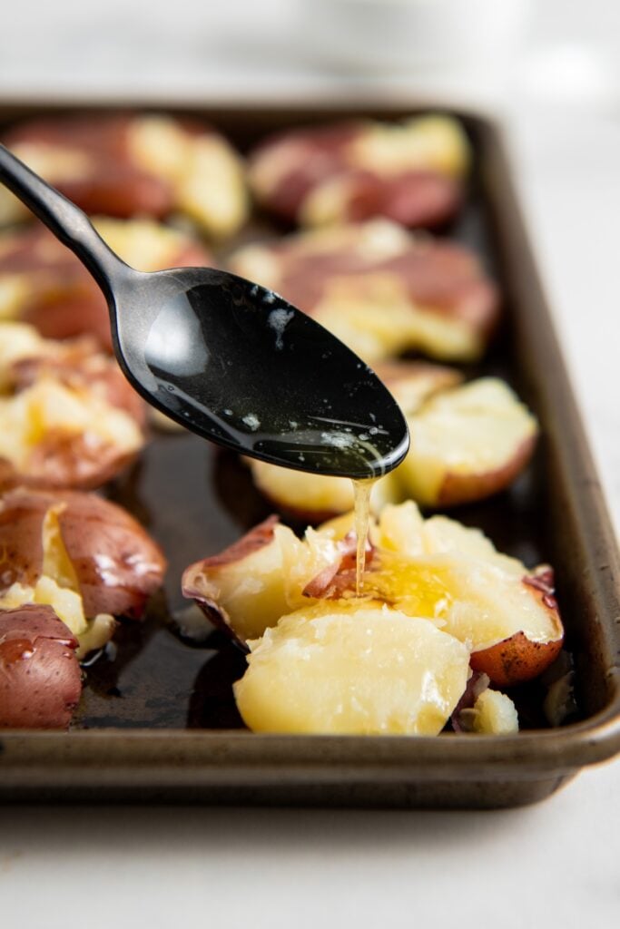 A black spoon drizzles olive oil over smashed red potatoes on a baking sheet, preparing them for roasting.