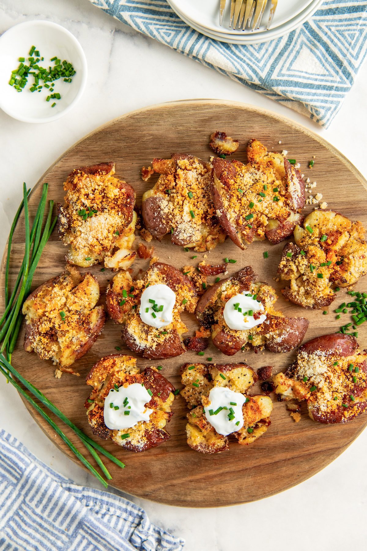 A round wooden board topped with smashed parmesan garlic roasted potatoes garnished with sour cream and chopped chives. Fresh chives and a bowl of chives are on the side, and a blue patterned napkin is nearby.