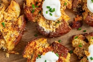 Crispy garlic parmesan smashed potatoes topped with sour cream, grated cheese, and chopped chives are arranged on a wooden board, with fresh chives placed nearby.