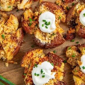 Crispy garlic parmesan smashed potatoes topped with sour cream, grated cheese, and chopped chives are arranged on a wooden board, with fresh chives placed nearby.