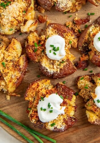 Crispy garlic parmesan smashed potatoes topped with sour cream, grated cheese, and chopped chives are arranged on a wooden board, with fresh chives placed nearby.