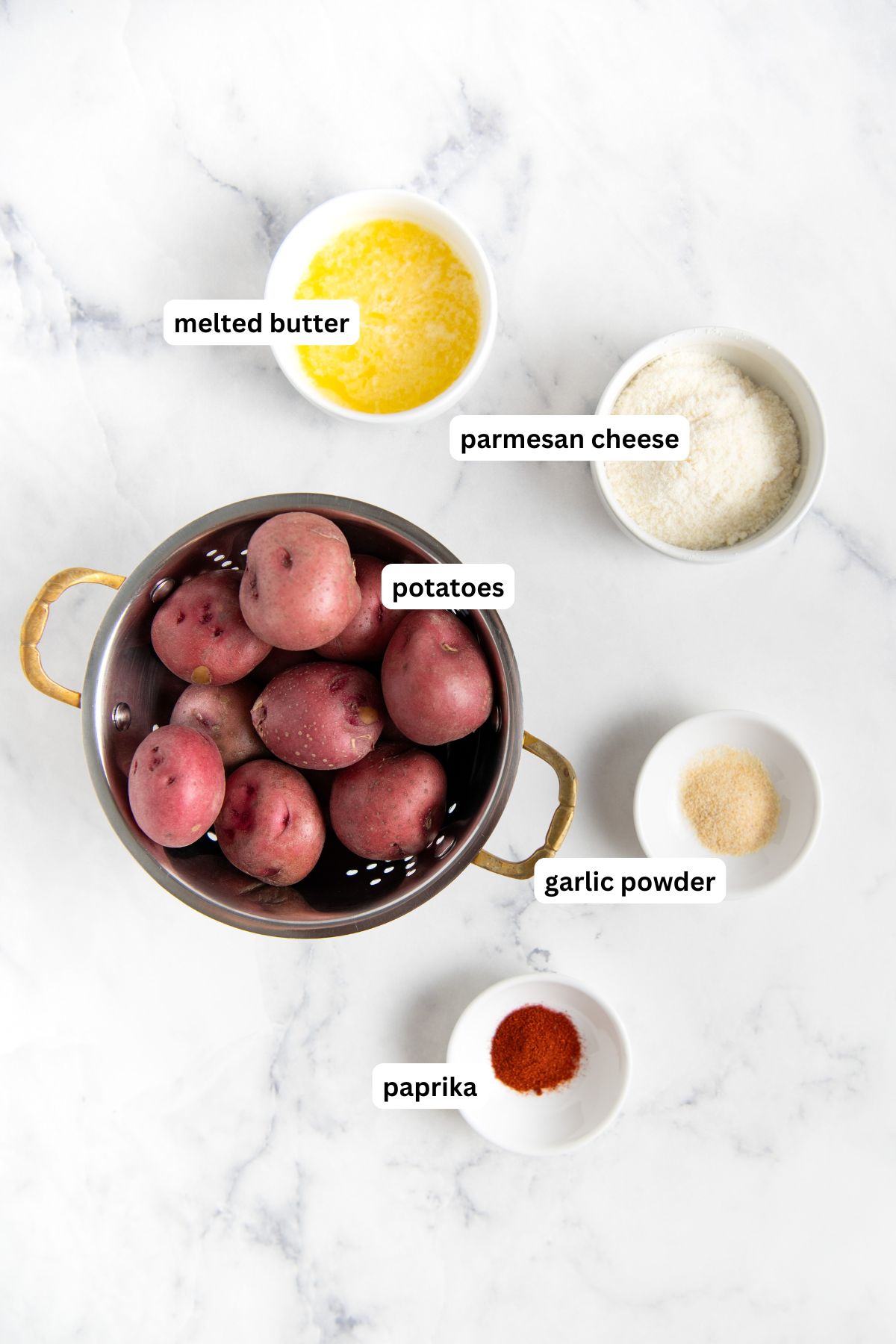 Ingredients for garlic parmesan smashed potatoes recipe arranged on a counter top. A bowl of red potatoes sits beside small bowls containing melted butter, grated parmesan cheese, garlic powder, and paprika, all labeled, on a white marble surface.