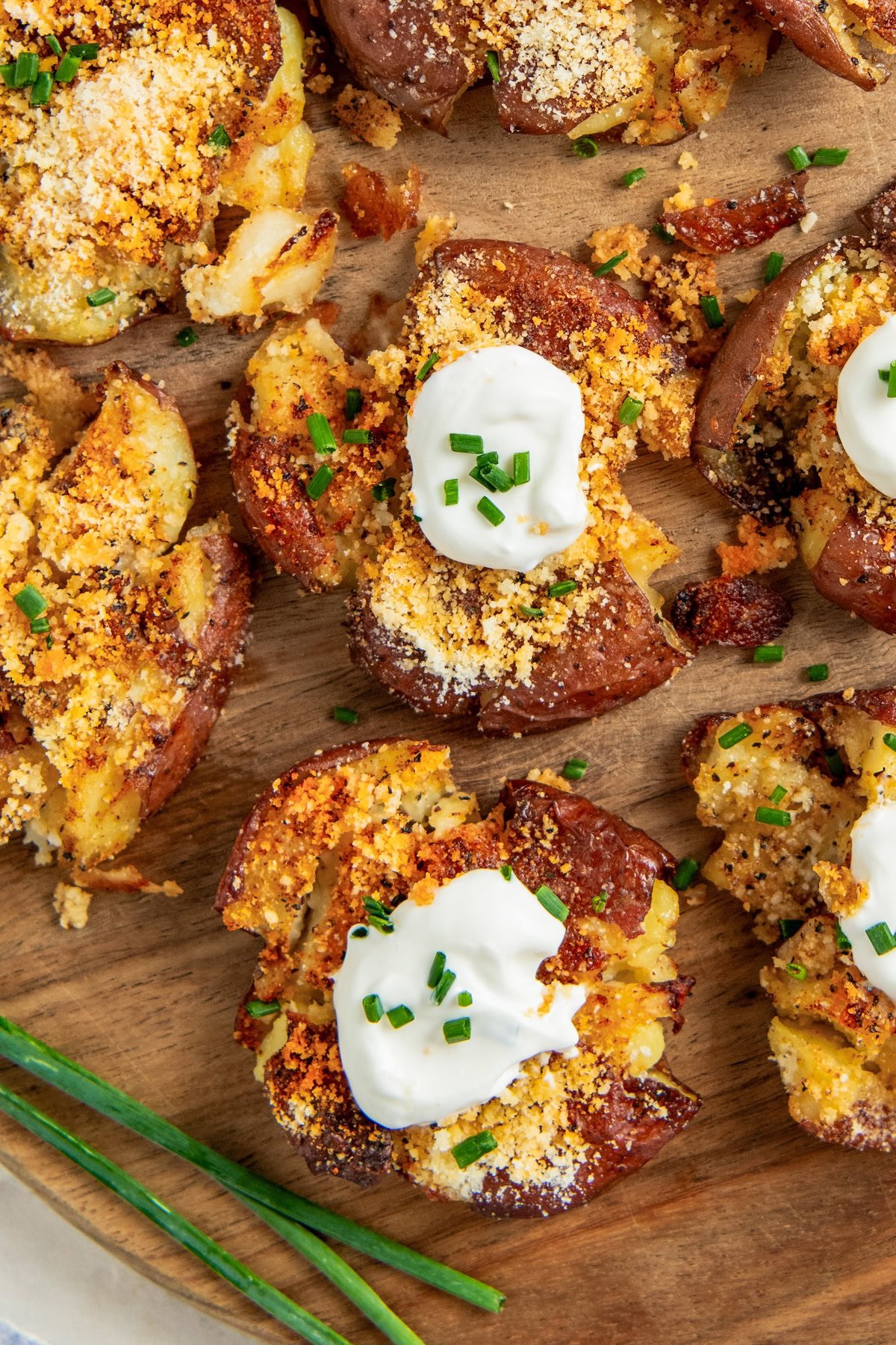 Crispy garlic parmesan smashed potatoes topped with sour cream, grated cheese, and chopped chives are arranged on a wooden board, with fresh chives placed nearby.
