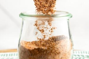 A measuring spoon is sprinkling a fajita seasoning mix into a glass jar, which is placed on a green and white patterned cloth.