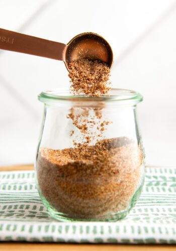 A measuring spoon is sprinkling a fajita seasoning mix into a glass jar, which is placed on a green and white patterned cloth.