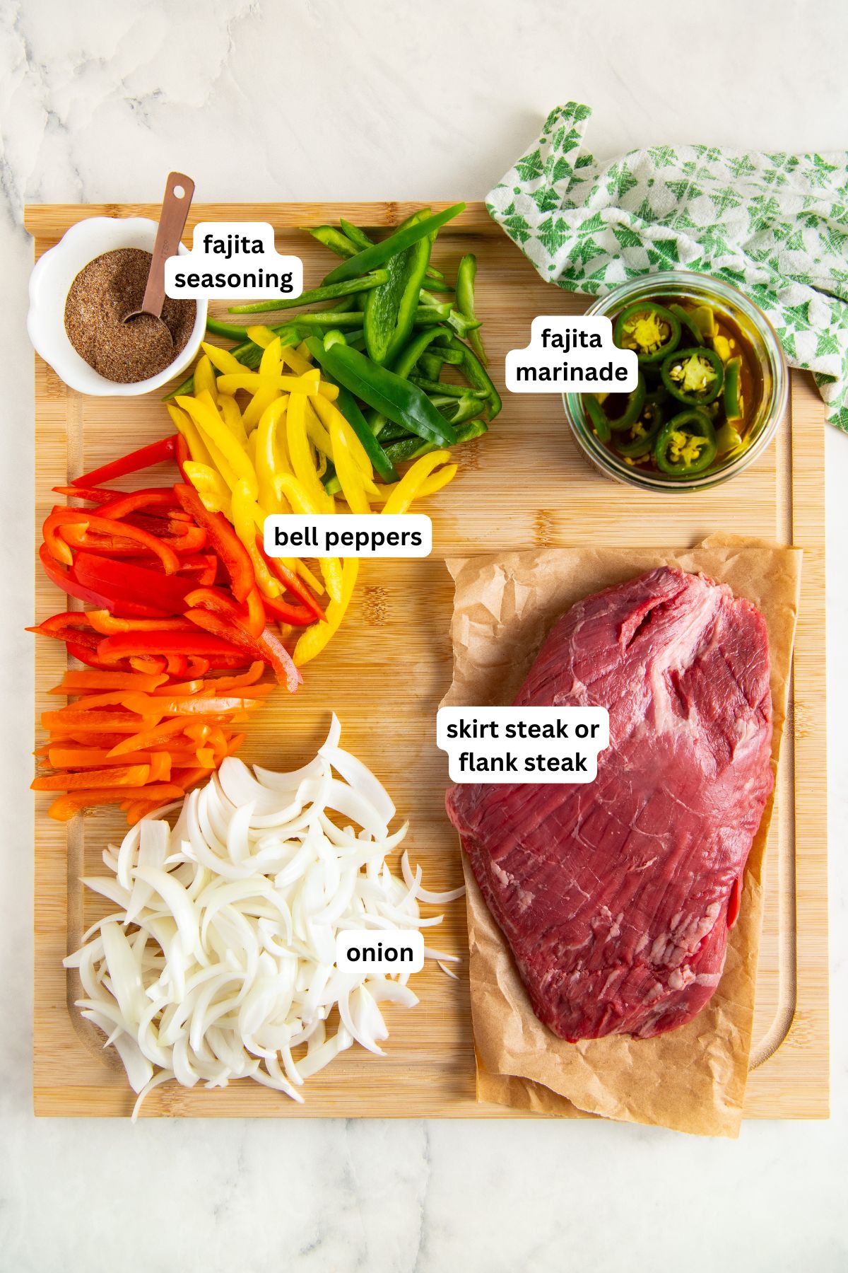 Ingredients for sheet pan fajita recipe arranged on a wooden board: sliced red, yellow, and green bell peppers, sliced onion, a raw skirt or flank steak, fajita seasoning in a small bowl, and a jar of fajita marinade with jalapeños.