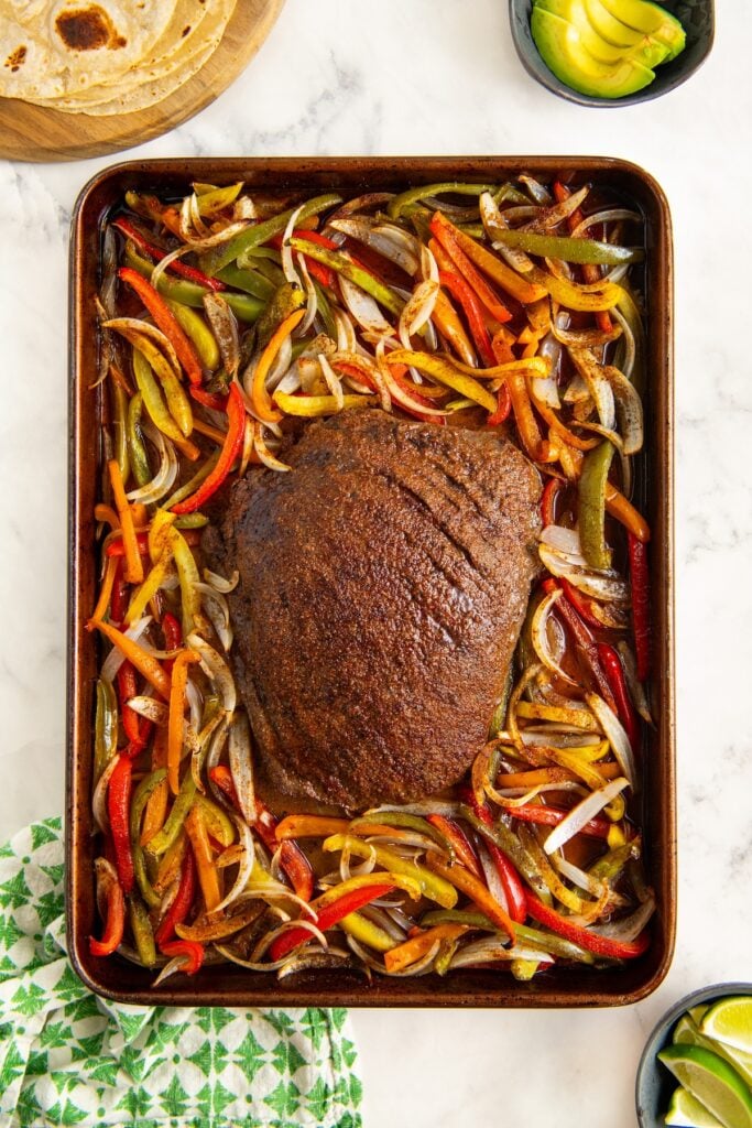 A sheet pan with a large, seasoned cooked fajita steak ready to be sliced, surrounded by sliced red, yellow, and green bell peppers and onions. Nearby are tortillas, sliced avocado, and lime wedges.