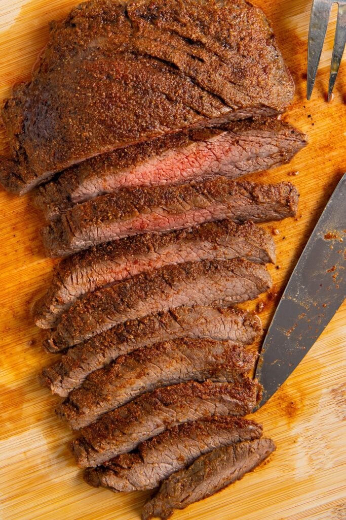 Sliced, seasoned fajita steak on a wooden cutting board, showing a pink center. A knife and meat fork are next to the slices.