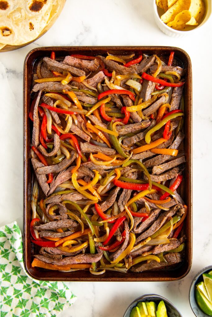 A sheet pan filled with cooked strips of beef fajitas, red, yellow, and green bell peppers, and onions. Bowls of tortilla chips, sliced avocado, and tortillas are nearby on a white countertop.
