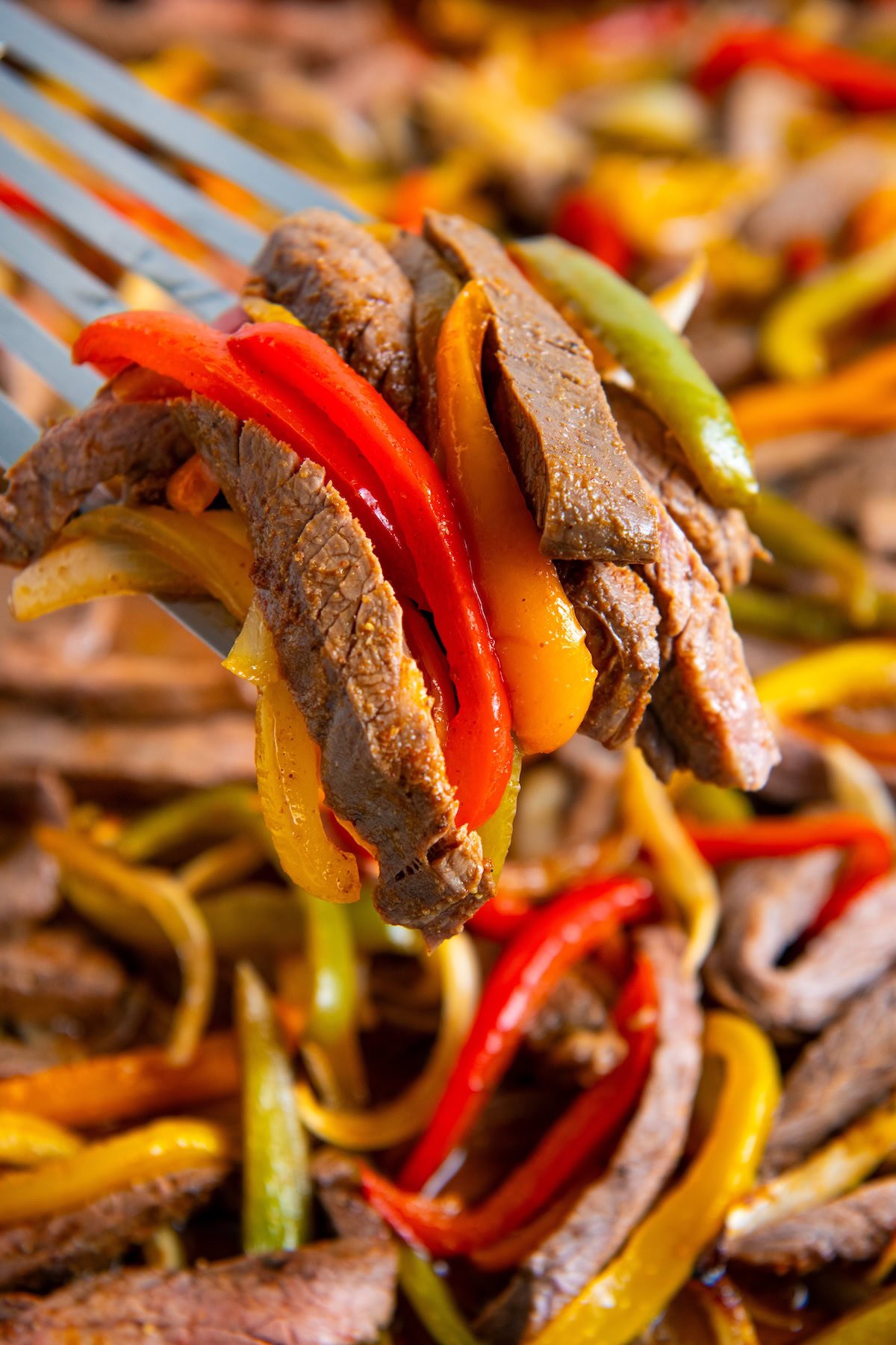 A close-up of a fork holding seasoned beef fajitas with red, yellow, and green bell peppers, with more beef and peppers blurred in the background.