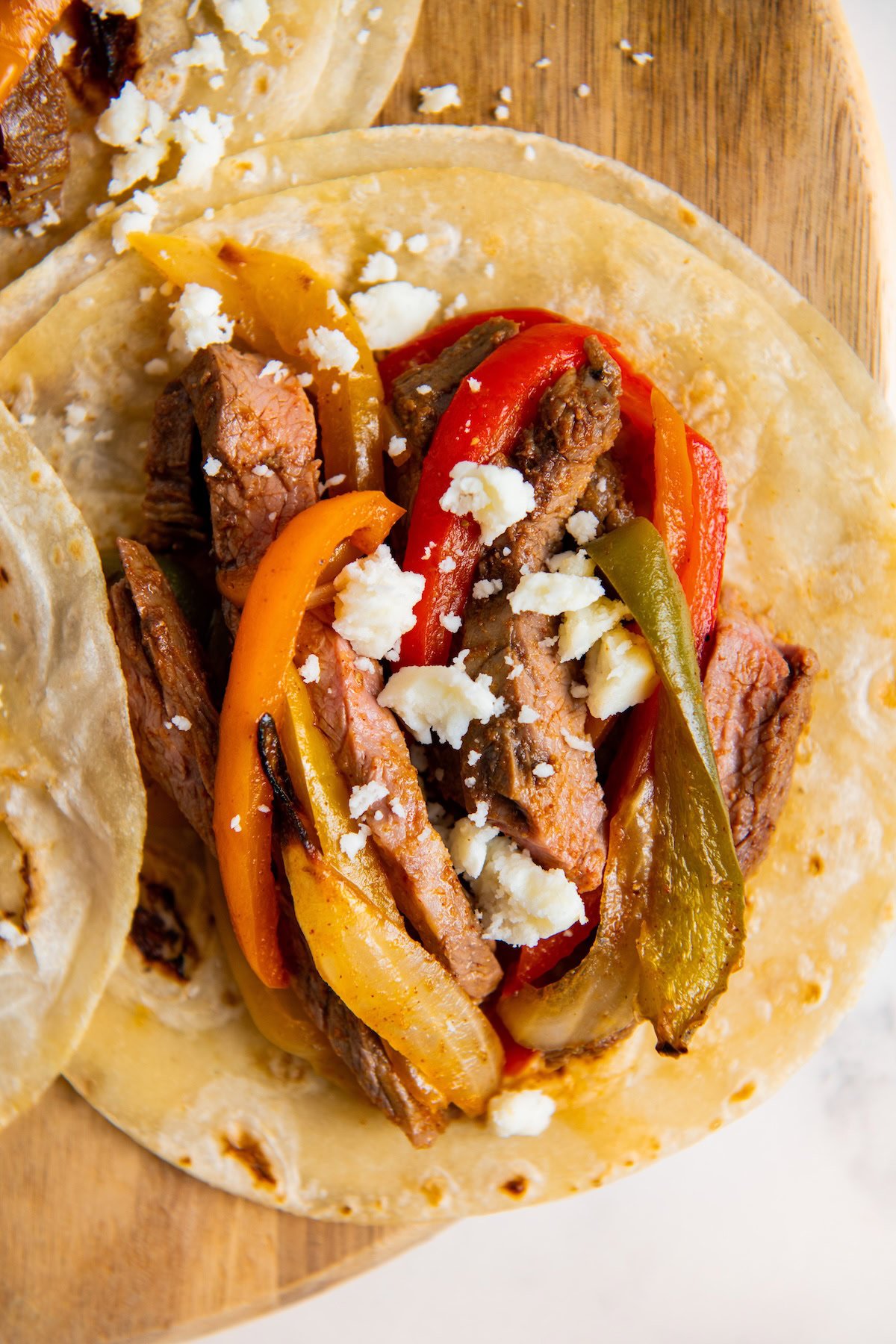 A close-up of a steak fajita taco on a wooden board, filled with grilled steak strips, sautéed red, yellow, and green bell peppers, onions, and topped with crumbled white cheese.