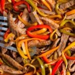 Sliced sheet pan steak fajitas mixed with red, yellow, and green bell peppers and onions, cooked and served on a tray with a metal spatula partially visible.