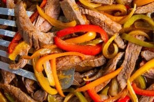 Sliced sheet pan steak fajitas mixed with red, yellow, and green bell peppers and onions, cooked and served on a tray with a metal spatula partially visible.