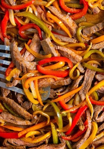 Sliced sheet pan steak fajitas mixed with red, yellow, and green bell peppers and onions, cooked and served on a tray with a metal spatula partially visible.