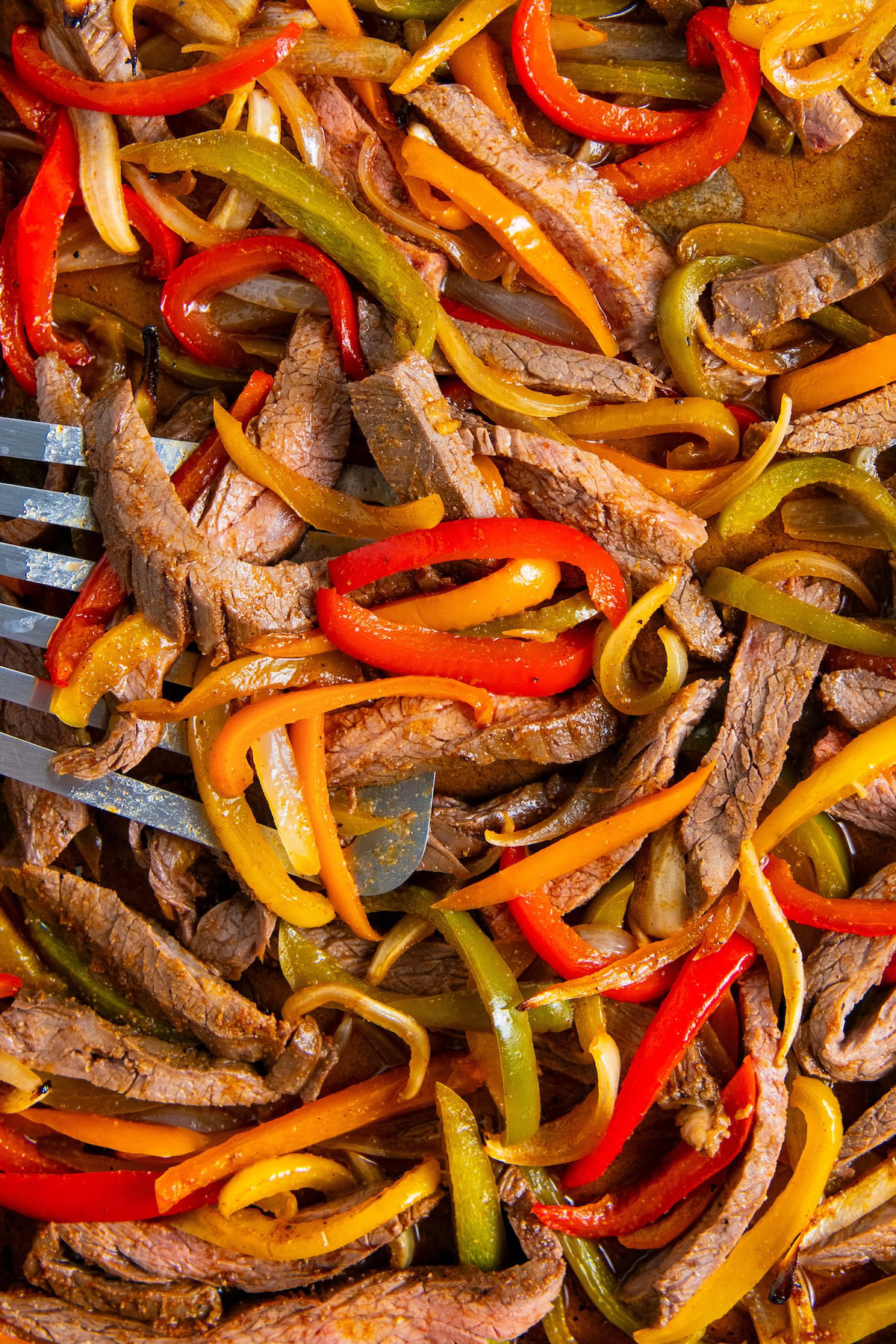 Perfect for busy weeknights, Sheet Pan Steak Fajitas feature juicy steak, colorful bell peppers, and sweet onions all roasted to perfection. Sliced sheet pan steak fajitas mixed with red, yellow, and green bell peppers and onions, cooked and served on a tray with a metal spatula partially visible.