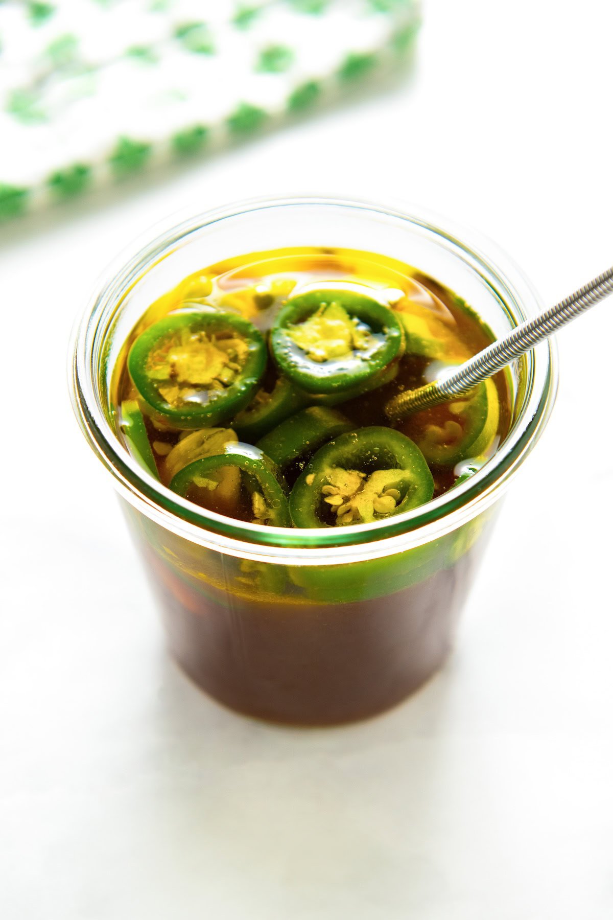 A glass jar filled with steak fajita marinade and topped with sliced green jalapeño peppers, with a metal spoon inside.