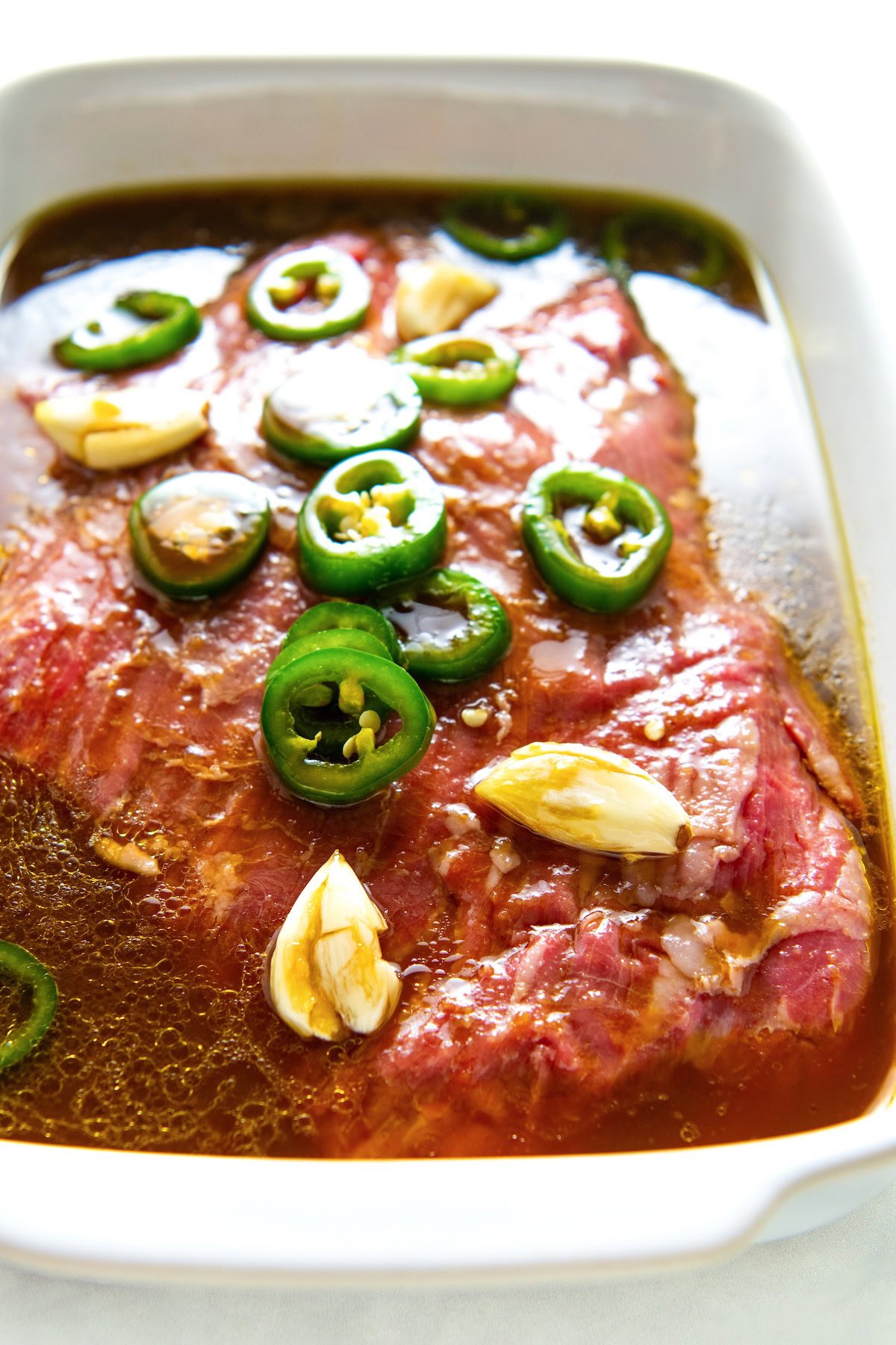 Raw beef in a white dish, topped with sliced jalapeños and whole garlic cloves, soaking in a fajita marinade.