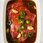 Raw beef marinating in a white dish with sliced jalapeños, garlic cloves, and a dark liquid steak fajita marinade on a white marble surface.