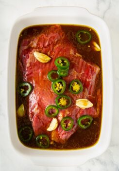 Raw beef marinating in a white dish with sliced jalapeños, garlic cloves, and a dark liquid steak fajita marinade on a white marble surface.
