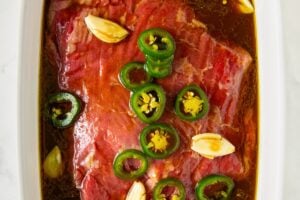 Raw beef marinating in a white dish with sliced jalapeños, garlic cloves, and a dark liquid steak fajita marinade on a white marble surface.