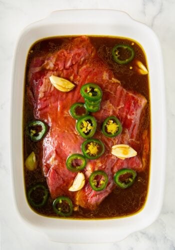Raw beef marinating in a white dish with sliced jalapeños, garlic cloves, and a dark liquid steak fajita marinade on a white marble surface.