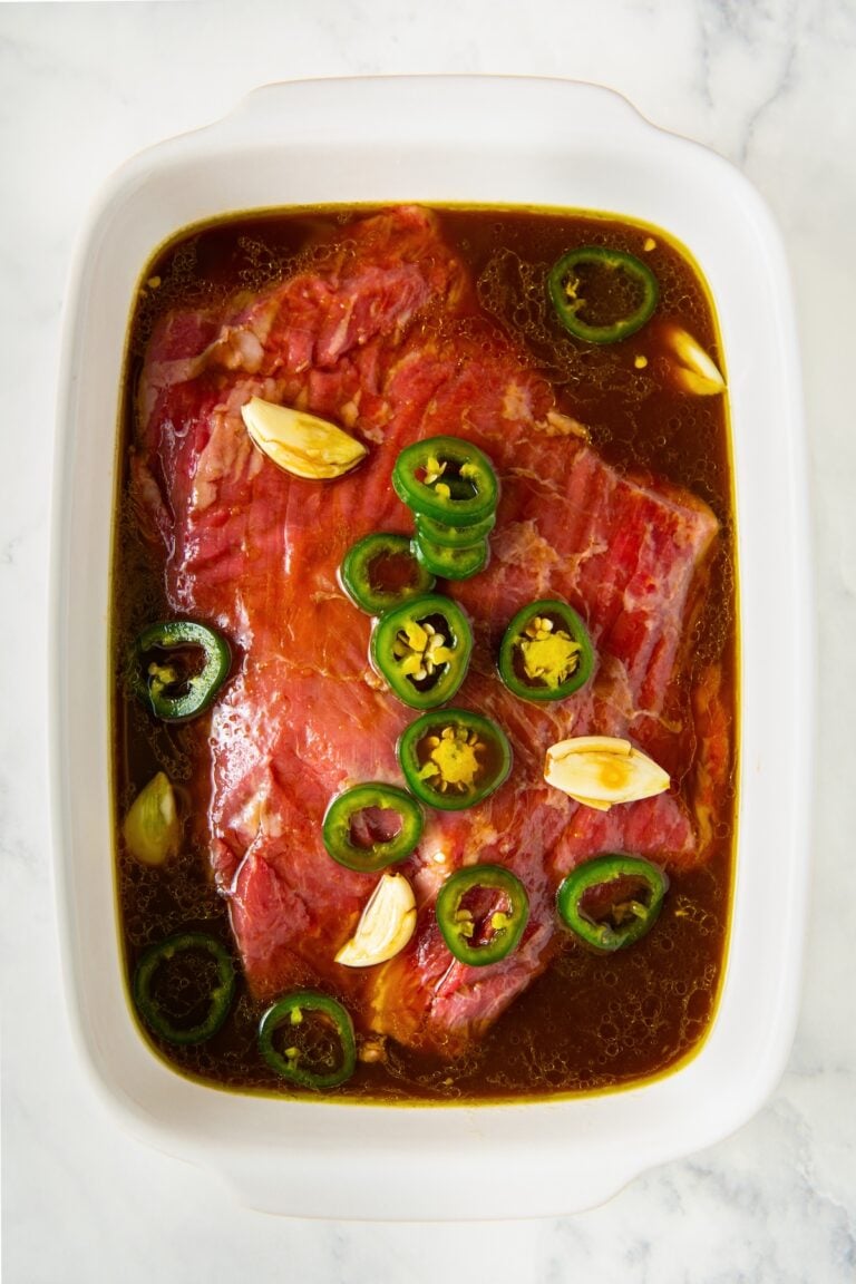 Raw beef marinating in a white dish with sliced jalapeños, garlic cloves, and a dark liquid steak fajita marinade on a white marble surface.