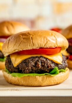 A close-up of a cheeseburger with a beef patty, cheddar cheese, tomato slice, and lettuce on a soft, golden bun. Two more burgers are blurred in the background.