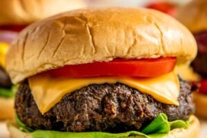 A close-up of a cheeseburger with a beef patty, cheddar cheese, tomato slice, and lettuce on a soft, golden bun. Two more burgers are blurred in the background.
