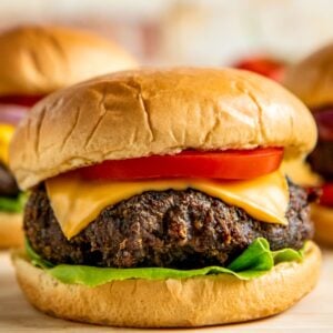 A close-up of a cheeseburger with a beef patty, cheddar cheese, tomato slice, and lettuce on a soft, golden bun. Two more burgers are blurred in the background.