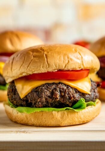 A close-up of a cheeseburger with a beef patty, cheddar cheese, tomato slice, and lettuce on a soft, golden bun. Two more burgers are blurred in the background.