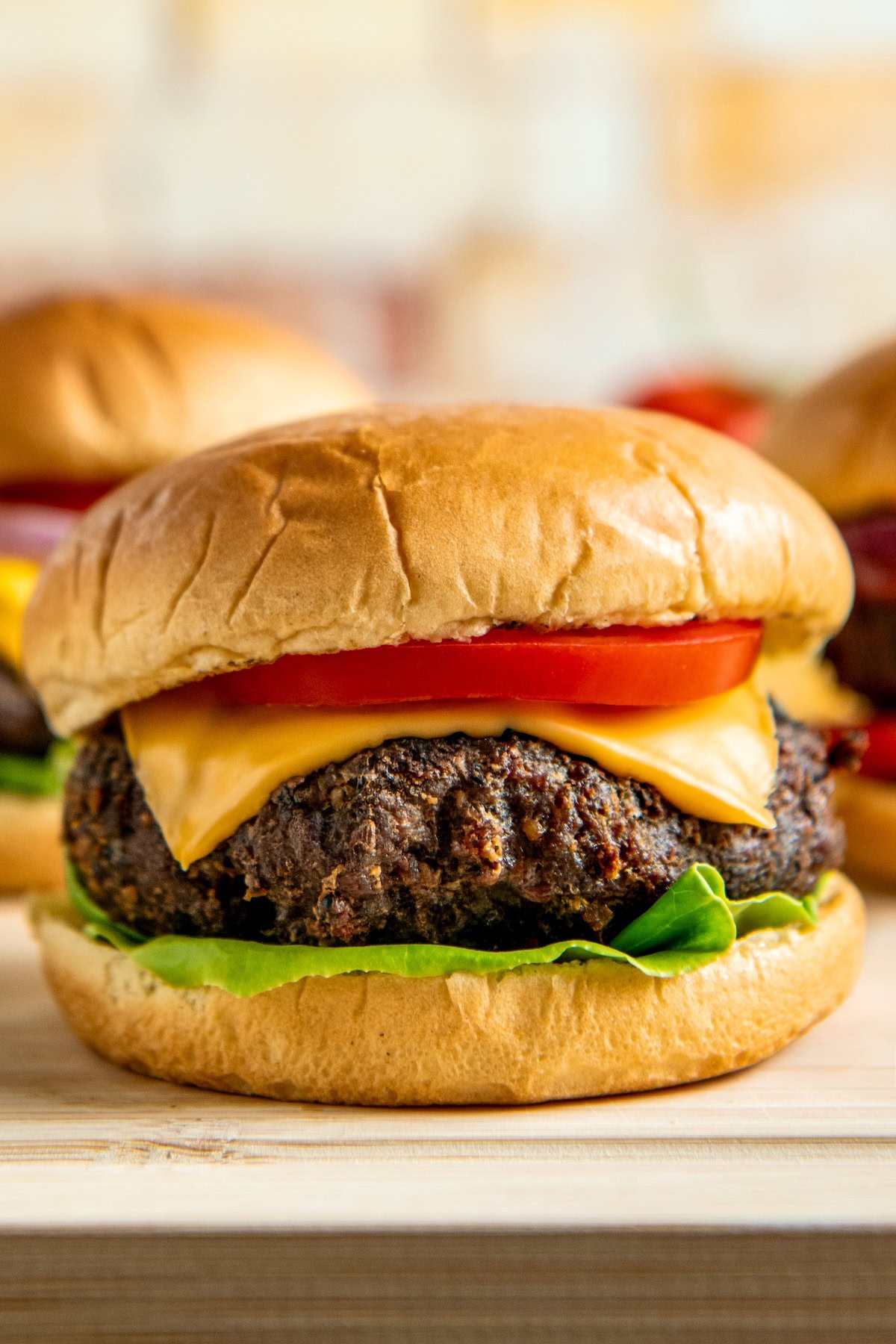 A close-up of a cheeseburger with a beef patty, cheddar cheese, tomato slice, and lettuce on a soft, golden bun. Two more burgers are blurred in the background.