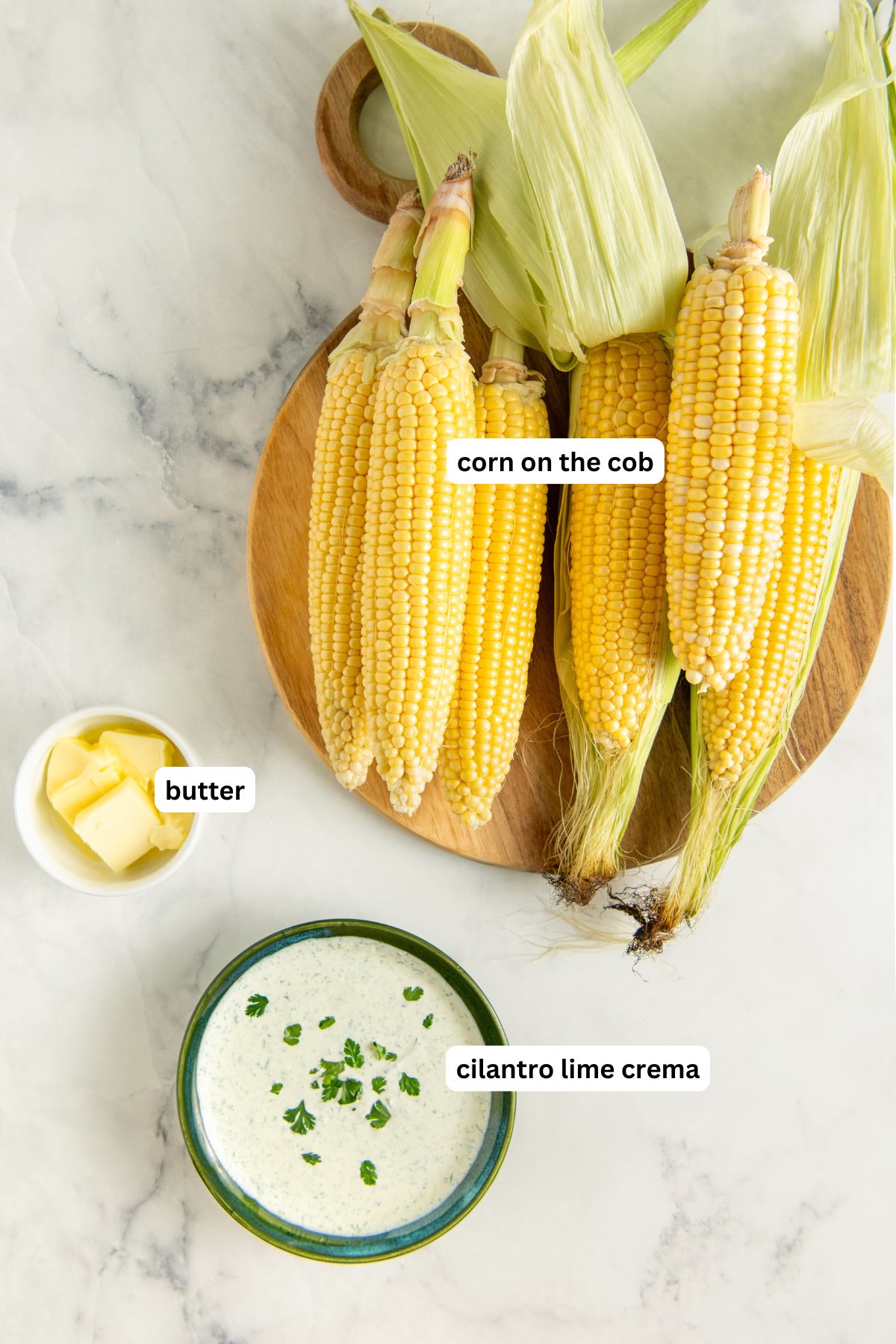 A wooden board with three ears of corn on the cob, a small bowl of butter cubes, and a bowl of cilantro lime crema garnished with chopped cilantro, all set on a light marble surface.