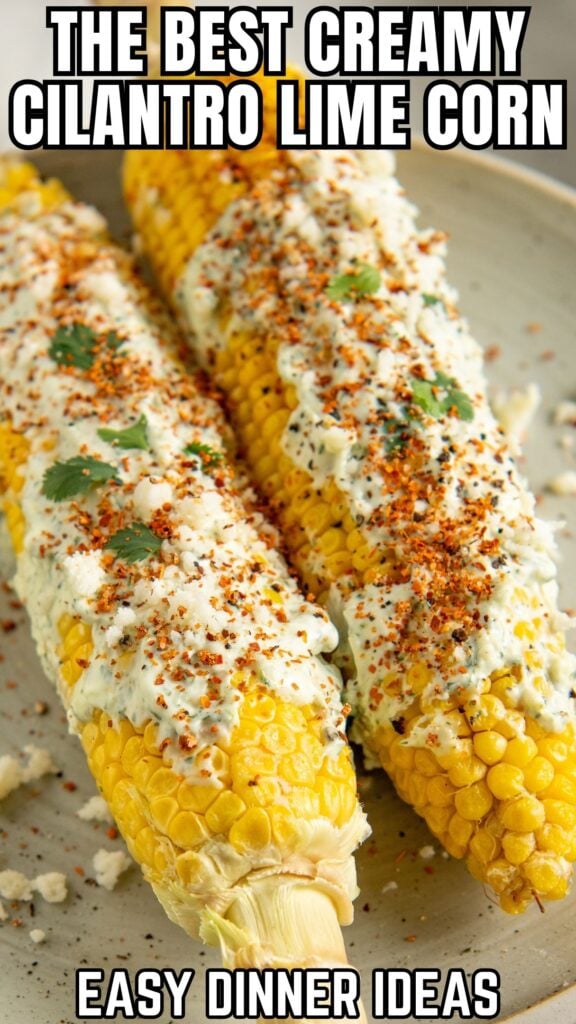 Two ears of Cilantro Lime Corn on the Cob topped with creamy cilantro lime sauce, chopped cilantro, crumbled mexican cheese, and tan, arranged on a plate.