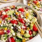 A bowl of fresh pearl couscous salad with cherry tomatoes, cucumbers, red onions, yellow bell peppers, olives, parsley, and crumbled feta cheese. Two plates with similar salad are in the background.