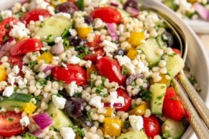 A bowl of fresh pearl couscous salad with cherry tomatoes, cucumbers, red onions, yellow bell peppers, olives, parsley, and crumbled feta cheese. Two plates with similar salad are in the background.