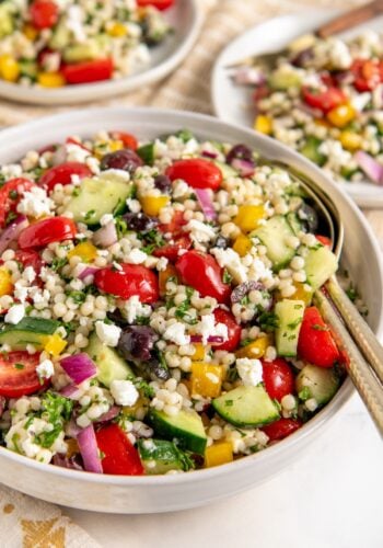 A bowl of fresh pearl couscous salad with cherry tomatoes, cucumbers, red onions, yellow bell peppers, olives, parsley, and crumbled feta cheese. Two plates with similar salad are in the background.