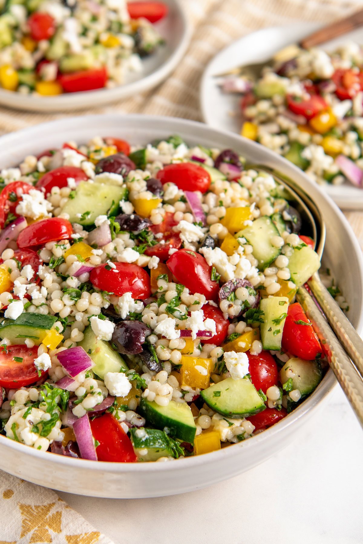 Mediterranean Pearl Couscous Salad with grape tomatoes, crisp cucumbers, salty olives & creamy feta all tossed in a zesty lemon vinaigrette. A bowl of fresh pearl couscous salad with cherry tomatoes, cucumbers, red onions, yellow bell peppers, olives, parsley, and crumbled feta cheese. Two plates with similar salad are in the background.