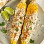 Two ears of cilantro lime corn on the cob covered in cilantro lime crema, sprinkled with crumbled cheese, tan, and cilantro, served on a plate with lime wedges and a few cilantro leaves.