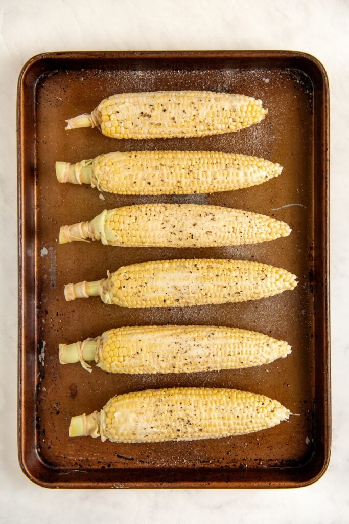 Six ears of corn, husked, are arranged in a single layer on a brown baking sheet. The corn is coated with butter and sprinkled with salt and black pepper, ready to be cooked.
