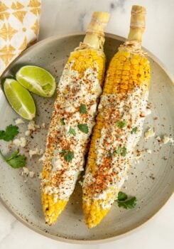 Two ears of cilantro lime corn on the cob covered in cilantro lime crema, sprinkled with crumbled cheese, tan, and cilantro, served on a plate with lime wedges and a few cilantro leaves.