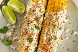 Two ears of cilantro lime corn on the cob covered in cilantro lime crema, sprinkled with crumbled cheese, tan, and cilantro, served on a plate with lime wedges and a few cilantro leaves.